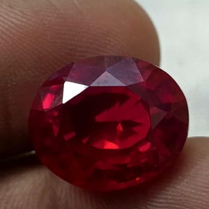 May include: A faceted, oval-shaped, deep red gemstone. The gem has multiple facets that reflect light, creating a brilliant sparkle. The stone is held between fingers, showcasing its size and color.