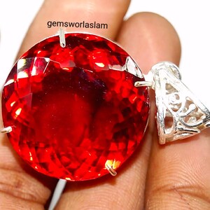 May include: A large, round, faceted red gemstone pendant set in a silver-toned metal frame with a decorative, filigree-style bail. The text "gemsworlaslam" is printed in black above the gemstone, which features a brilliant, reflective surface.