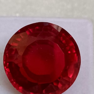 Op de afbeelding: Een gefacetteerde, ronde, dieprode edelsteen. De edelsteen heeft een briljante slijpvorm met vele facetten die het licht reflecteren. De kleur is een rijk, verzadigd rood, met een hoge mate van helderheid. De edelsteen is waarschijnlijk een robijn.