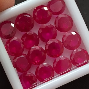 Puede incluir: Una colección de rubíes redondos, facetados, de color rojo intenso, exhibidos en una bandeja de plástico blanco. Las piedras preciosas tienen un alto brillo y están dispuestas muy juntas, mostrando su vibrante color y claridad. La bandeja está ligeramente inclinada.