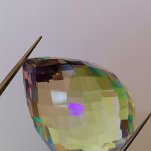 Pode incluir: Uma gema facetada em forma de pêra com uma mistura de cores, incluindo amarelo, verde, roxo e azul. A gema é segurada por duas pinças de metal. A superfície da gema reflete a luz, criando um efeito arco-íris.