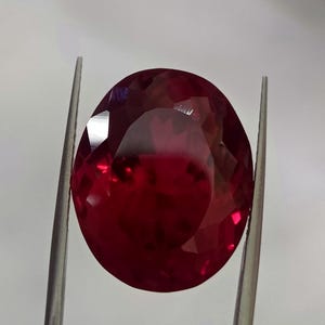 Può includere: Una gemma di rubino sfaccettata, di forma ovale, tenuta da una pinzetta su sfondo bianco. Il rubino è di un colore rosso intenso e saturo, con molteplici sfaccettature che catturano la luce. La pinzetta è argentata.