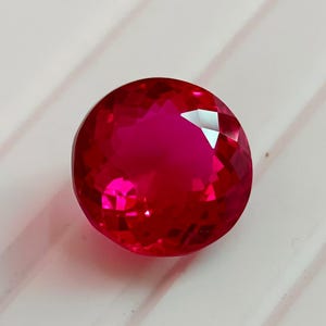 May include: A round, faceted gemstone with a vibrant, deep red colour. The stone features a brilliant cut with numerous triangular and kite-shaped facets that reflect light, creating a sparkling appearance across its circular surface.
