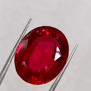 Puede incluir: Un rubí de talla ovalada, de color rojo intenso, sujeto con pinzas plateadas. La gema presenta facetas internas que reflejan la luz. La piedra preciosa se encuentra sobre un fondo blanco liso, resaltando su color y claridad.