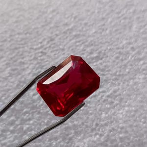 Può includere: Una pietra preziosa di colore rosso intenso con taglio rettangolare, tenuta da una pinzetta su uno sfondo bianco.