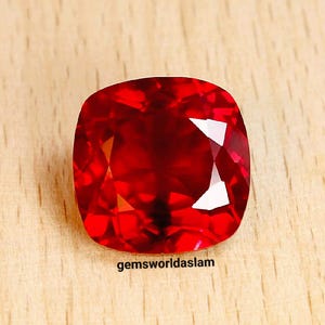 May include: A deep red gemstone with a cushion cut. The gemstone is on a wooden surface. The text "gemsworldaslam" is visible below the gemstone.