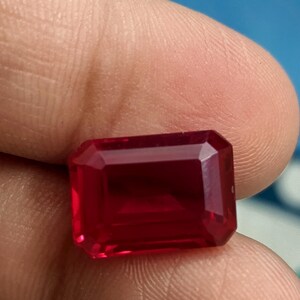 Op de afbeelding: Een dieprode edelsteen met een rechthoekige smaragdslijp. De edelsteen wordt in een hand gehouden.