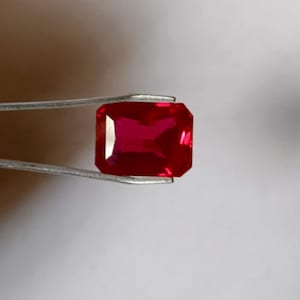 Könnte beinhalten: Ein tief roter Edelstein mit einem rechteckigen Smaragdschliff, in einer Pinzette vor einem weißen Hintergrund gehalten.