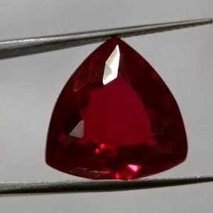 Puede incluir: Una gema de rubí facetada en forma triangular. El rubí es de un color rojo intenso y profundo, sostenido por unas pinzas. La gema tiene múltiples facetas que reflejan la luz, creando un efecto brillante. Adecuado para joyería.