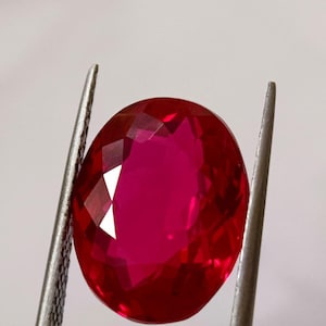 May include: A deep red, oval-cut ruby gemstone held by silver tweezers. The gemstone's facets reflect light, creating a sparkling effect. The background is plain white.