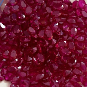 May include: A pile of faceted, oval-shaped rubies. The gemstones are a deep, rich red color, with multiple facets catching the light. The rubies are scattered, creating a dense, textured appearance. The image is a close-up, highlighting the clarity and brilliance of the stones.