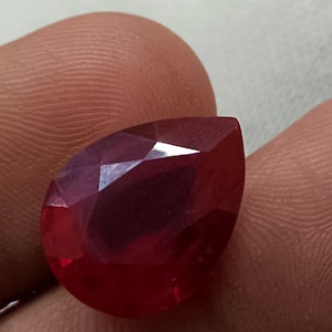 Pode incluir: Uma gema de rubi facetada em forma de pêra. A gema é de uma cor vermelha profunda e rica, com múltiplas facetas que capturam a luz. O rubi é segurado entre os dedos, mostrando seu tamanho e forma.