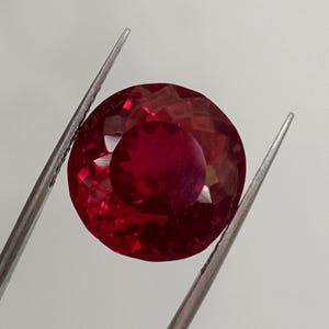 May include: A round-cut ruby gemstone, held by silver tweezers. The ruby is a deep red colour, with multiple facets reflecting light. The tweezers hold the gemstone against a plain white background.