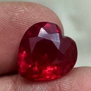 May include: A faceted, heart-shaped ruby gemstone. The deep red stone has multiple facets that reflect light, creating a sparkling effect. The gemstone is held between fingers.