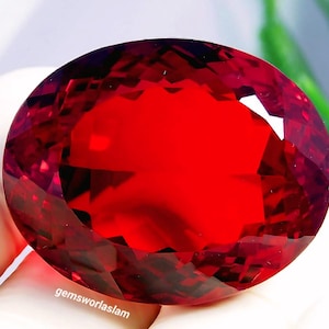 May include: A large, oval-cut, vibrant red gemstone with a faceted surface that reflects light. The stone features a deep crimson hue and is marked with the text "gemsworlaslam" in small white letters along the lower left edge of the facet.