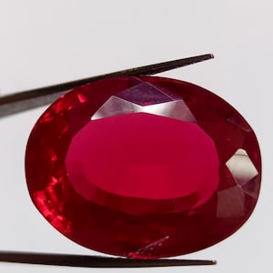 Puede incluir: Un rubí facetado de forma ovalada, sujeto con unas pinzas. El rubí es de color rojo intenso y saturado, con una superficie pulida. La gema se encuentra sobre un fondo blanco liso.
