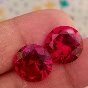 Può includere: Due pietre preziose rotonde e sfaccettate di colore rosso. Le pietre preziose sono approssimativamente della stessa dimensione e hanno una finitura lucida e lucidata.