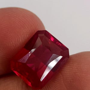 May include: A faceted, rectangular-cut ruby gemstone. The gem is a deep, saturated red color with multiple facets that reflect light. The gemstone is held between two fingers.