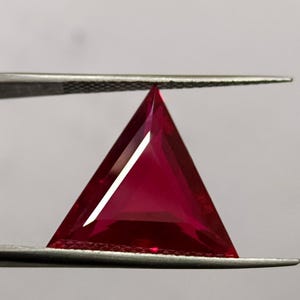 May include: A triangular, faceted, deep red gemstone is held by tweezers against a white background. The gem has sharp edges and a polished surface, reflecting light. The shape is a perfect equilateral triangle.