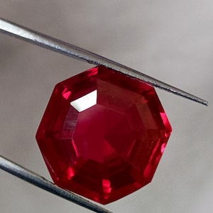 May include: A faceted, octagonal, deep red gemstone is held by a pair of silver tweezers. The gem has a brilliant, reflective surface, showcasing its many facets. The gemstone's color is a rich, saturated red.