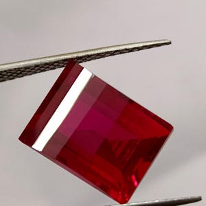 May include: A faceted, rectangular ruby gemstone held by tweezers. The gem is a deep, vibrant red with sharp, clean edges and a reflective surface. The gemstone is held against a neutral background.
