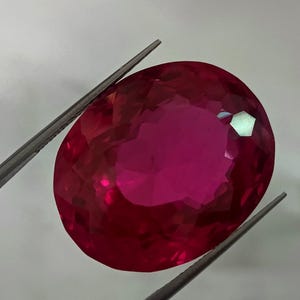 May include: A large, oval-shaped, faceted gemstone with a deep pink hue. The gemstone is held in place by a pair of tweezers.