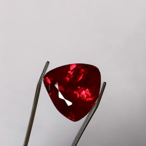 Pode incluir: Uma gema vermelha escura segurada por pinças. A gema tem a forma de um coração com facetas.