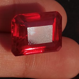Pode incluir: Uma gema vermelha escura com um corte quadrado esmeralda. A gema está engastada em uma moldura de prata.