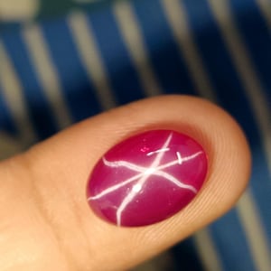 Pode incluir: Gema ovalada, vermelho escuro, com um proeminente efeito de estrela de seis raios. Linhas brancas se cruzam sobre a superfície polida. O fundo está desfocado.