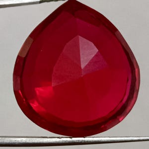 May include: A faceted, teardrop-shaped gemstone in a vibrant red hue. The gem is held by silver tweezers, showcasing its polished surface and internal facets. The gem's shape and color suggest it could be a ruby or a similar precious stone.