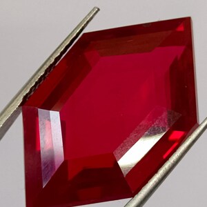 May include: A faceted, deep red gemstone held by silver tweezers. The gem has a unique, geometric shape with multiple facets that reflect light. The color is a rich, vibrant red, and the gem appears to be polished.