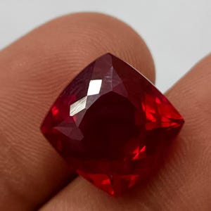 Op de afbeelding: Een gefacetteerde robijn edelsteen met kussensnede. De edelsteen heeft een diepe, verzadigde rode kleur met briljante facetten die het licht reflecteren. De edelsteen wordt tussen de vingers gehouden, wat de grootte en helderheid aantoont.