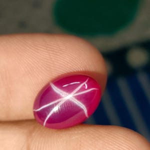May include: Oval-shaped, polished, ruby gemstone with a six-rayed star effect. The gem is a deep, rich pinkish-red color with white star lines. The stone is held between fingers, showcasing its size and clarity.