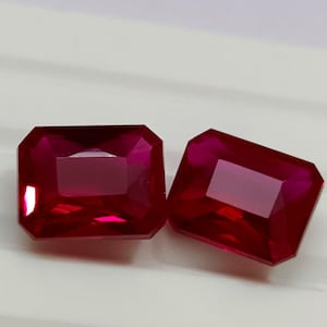 Può includere: Due gemme rosso intenso con taglio a raggiera. Le gemme hanno una forma rettangolare con bordi smussati, che riflettono la luce e mettono in risalto il loro colore intenso. Le pietre sono su uno sfondo bianco.