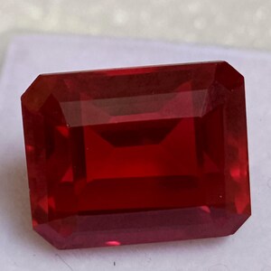 May include: A rectangular-cut, deep red gemstone with multiple facets. The gem has a clear, transparent appearance with sharp edges and a polished surface. The colour is a rich, saturated red. The gemstone is set against a neutral background.