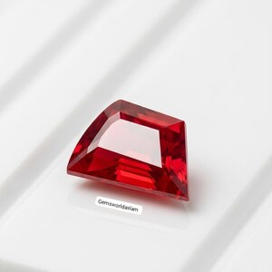 Può includere: Una gemma rossa vibrante con sfaccettature trapezoidali e superficie lucida. La pietra presenta bordi netti e puliti con una chiarezza brillante e riflettente. Una piccola etichetta bianca con il testo nero Gemsworldaslam si trova sotto.