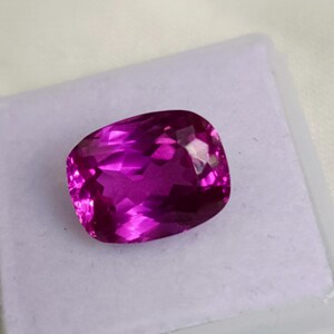 May include: A faceted, cushion-cut gemstone in a vibrant magenta hue. The gem is set against a white background, showcasing its clarity and brilliance. The gemstone is likely a ruby or a similar precious stone.