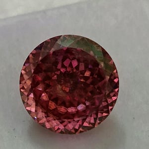 May include: A faceted, round gemstone with a deep pink hue, transitioning to lighter shades towards the edges. The gem displays a complex internal structure, reflecting light and creating a sparkling effect. The stone is set against a neutral background.
