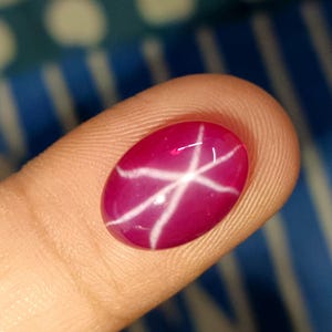 May include: A polished, oval-shaped, ruby-colored gemstone with a six-rayed star pattern. The star is white and appears to float within the gem. The gemstone is held between two fingers, showcasing its vibrant color and unique optical effect.