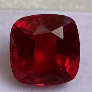 May include: A faceted, cushion-cut gemstone with a deep red hue. The gem's surface reflects light, showcasing its multiple facets and internal structure. The shape is square with rounded corners, typical of a cushion cut.