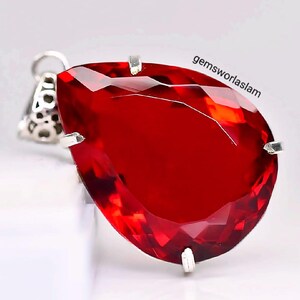 May include: A pear-shaped, faceted red gemstone pendant set in a silver-toned metal prong setting. The pendant features a decorative filigree bail at the top. The text gemsworlaslam is printed in black on the upper right side of the red stone.