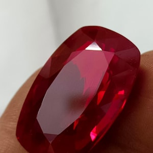 Pode incluir: Uma grande pedra preciosa de rubi facetada em forma retangular. A gema é de uma cor vermelha profunda e rica com múltiplas facetas que refletem a luz. A pedra preciosa é segurada em uma mão.