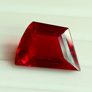 Op de afbeelding: Een gefacetteerde, dieprode edelsteen met een unieke, geometrische vorm. De edelsteen heeft een gepolijst oppervlak dat licht reflecteert, waardoor de helderheid en schittering worden benadrukt. De slijpvorm heeft een gebogen rand en meerdere facetten, wat een opvallend visueel effect creëert. De edelsteen ligt op een witte achtergrond.