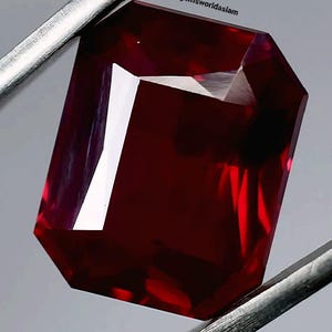 May include: A deep red gemstone with a rectangular cut and a polished finish. The gemstone is held by a pair of tweezers.