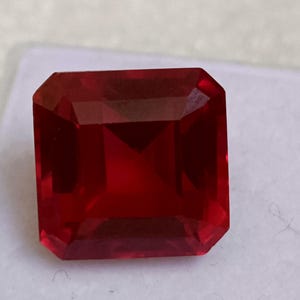 以下が含まれることがあります： 長方形にカットされ、ファセット加工された、深い赤色の宝石。宝石は、光を反射する複数のファセットを持つ対称的な形状をしています。色は豊かで飽和した赤色で、宝石は透明に見えます。