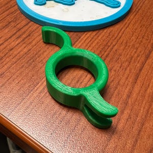 May include: A bright green plastic clip with a central circular opening and two curved, opposing arms. Positioned behind it is a circular white coaster featuring a raised blue lion logo with a blue border, showcasing a distinct 3D printed texture.
