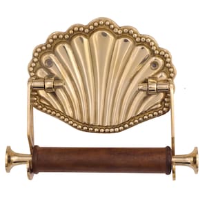 May include: A decorative brass toilet paper holder with a seashell design. The holder features a fan-shaped backplate with a beaded border and a wooden roller. The brass has a polished, golden finish.