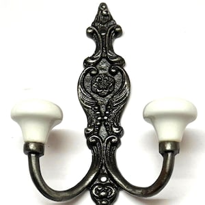 Puede incluir: Un colgador de pared de doble gancho con un diseño de metal negro adornado y dos pomos de cerámica blanca.