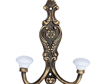 May include: Ornate, antique-style wall hook with a bronze finish and two curved arms. The hook features a decorative floral design and white ceramic knobs. Ideal for hanging coats, hats, or other items.