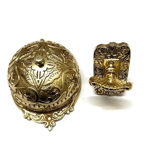 May include: A gold-toned doorbell with an ornate design. The doorbell has a round bell and a separate push button. The bell and button are both decorated with intricate floral patterns.